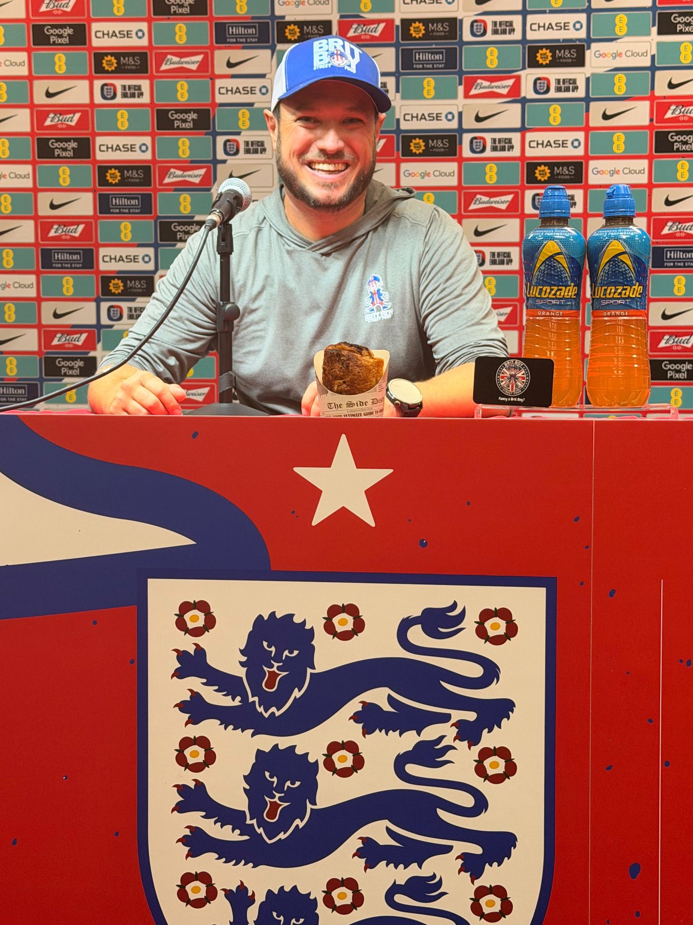 Chris Thomas at the England press conference podium in Kansas City, holding a Brit Boy sausage roll